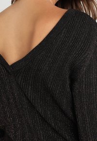 Svart ribbad tröja med djup V-ringning och metalltråd accenter, som visar en texturerad yta och en avslappnad silhuett.