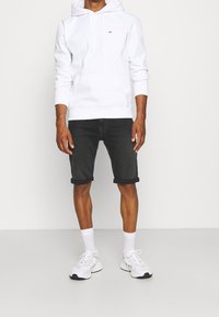 Sudadera blanca con bolsillo frontal y pequeño logo, combinada con pantalones cortos de mezclilla negros enrollados en el dobladillo. Zapatillas blancas y calcetines tobilleros completan el look.