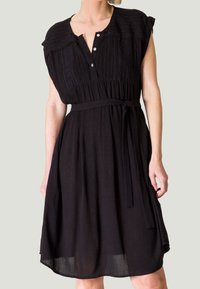 Robe noire à manches courtes, avec une patte de boutonnage, un tissu texturé et une ceinture nouée à la taille. Jupe fluide qui tombe juste au-dessus des genoux.