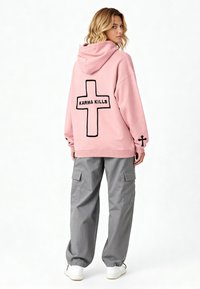 Hoodie rosa con grafica a croce nera e testo "KARMA KILLS" sul retro, abbinata a pantaloni cargo grigi e sneakers bianche.