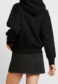 Persona con una sudadera negra y una minifalda de mezclilla gris oscura, vista por detrás con una mano relajada y la otra ligeramente doblada.