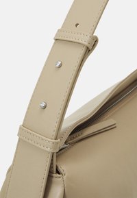 Cinghia di pelle beige con doppie borchie in argento impresse con "ck" vicino alla cerniera di una borsa in pelle beige coordinata.