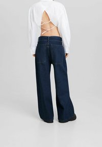 Vit långärmad skjorta med öppen ryggdesign och banddetaljer, matchad med djupblå vida jeans med två bakfickor.