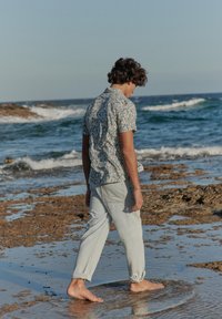 Joven con cabello rizado que lleva una camisa estampada y pantalones claros caminando descalzo por una costa rocosa con olas de fondo.