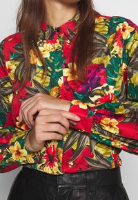 Chemise à boutons rouge à motif floral avec des motifs jaunes et verts vibrants, manches froncées et col classique, associée à du cuir noir.