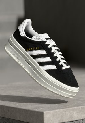 Czarno-biały sneaker Adidas Gazelle z białymi sznurowadłami i złotymi literami, pokazany unoszący się nad betonową powierzchnią.
