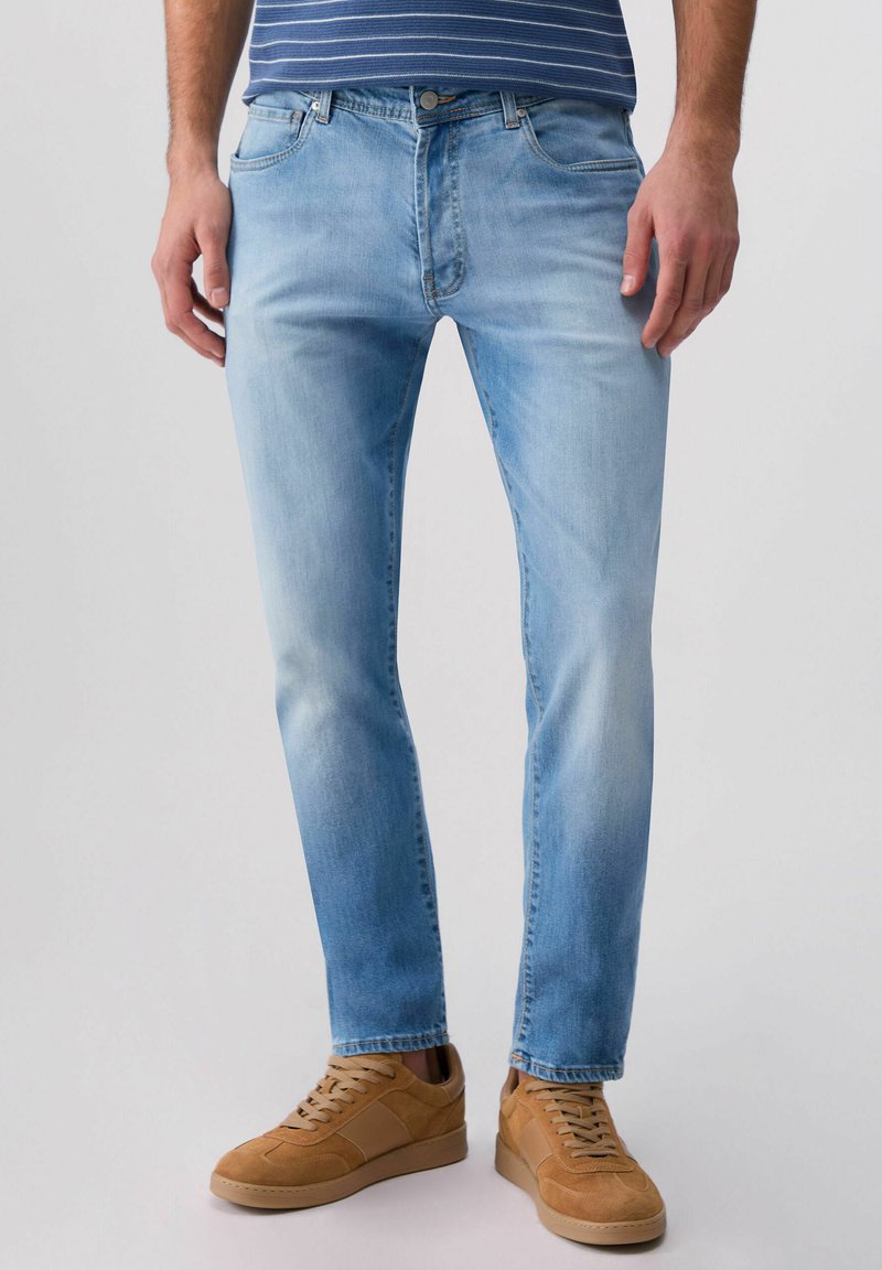 Bărbat purtând blugi slim fit de culoare bleu deschis, adidași bej din piele întoarsă și o cămașă albastră cu dungi, stând în picioare pe un fundal simplu.