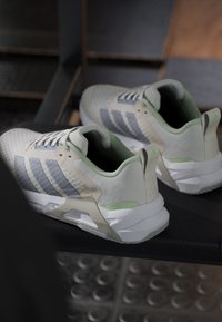 Бели и сиви атлетични обувки Adidas със зелени акценти, поставени върху фитнес оборудване с видим метален под под тях.