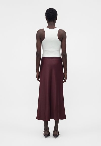 Γυναίκα που στέκεται με την πλάτη γυρισμένη φορώντας λευκό αμάνικο ριμπ μπλουζάκι, μακριά βουργουνδί φούστα και σκούρα πέδιλα με τακούνι σε λευκό φόντο.