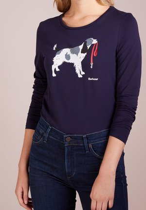 Vrouw draagt een marineblauw shirt met lange mouwen met een afbeelding van een witte en grijze hond die een rood riempje vasthoudt op de voorkant, gecombineerd met donkerblauwe spijkerbroek.