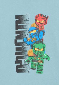 Helles blaues T-Shirt mit drei Cartoon-Ninja-Charakteren in rot, blau und grün. Fettschwarzer Text zeigt "Ninjago" neben ihnen.