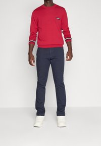 Sweat-shirt rouge avec accents noirs et blancs, col rond et manches longues, associé à un pantalon bleu marine et des baskets blanches.