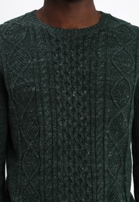 Dunkelgrüner Strickpullover mit strukturierten Mustern, darunter Zöpfe und Rauten, im klassischen Rundhalsausschnitt-Design.