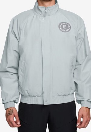 Homme portant une veste zippée gris clair avec un logo rond sur la poitrine gauche et un pantalon noir, debout, les mains détendues le long du corps.