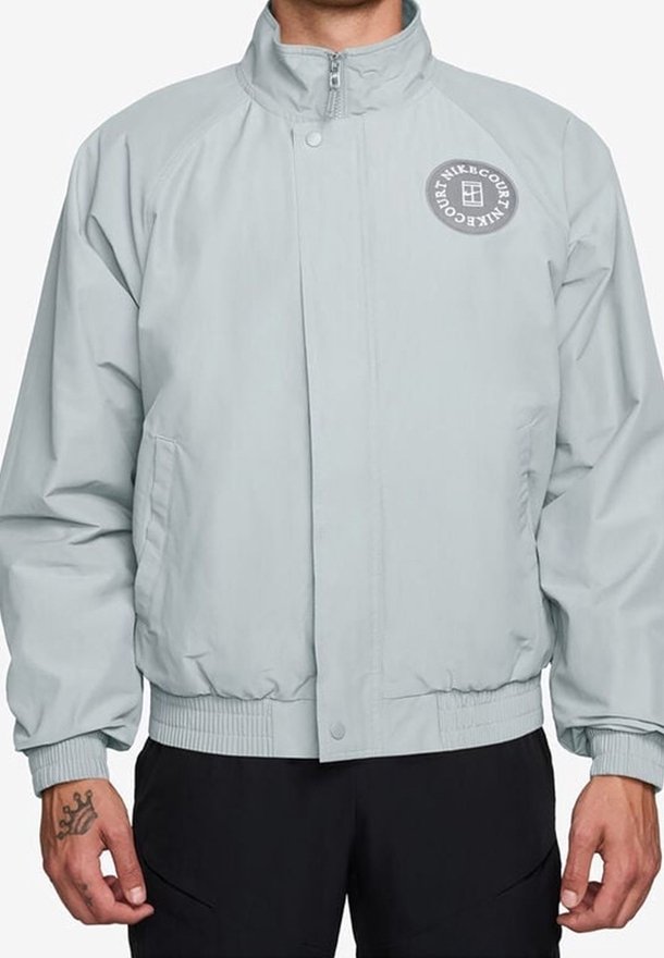 Homme portant une veste zippée gris clair avec un logo rond sur la poitrine gauche et un pantalon noir, debout, les mains détendues le long du corps.