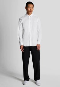 Camicia bianca a maniche lunghe con bottoni, realizzata in tessuto, indossata con pantaloni neri e sneakers bianche; presenta un colletto e un leggero dettaglio del logo.