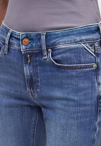 Gros plan sur un jean en denim bleu à la texture délavée, bouton en cuivre et étiquette « REPLAY » sur la poche avant, associé à un top gris côtelé.