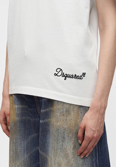 Άτομο με λευκή αμάνικη μπλούζα Dsquared2 με μαύρο λογότυπο κοντά στο στρίφωμα και φαρδιά τζιν μπλε με ξεθωριασμένο look, εμφανίζεται από τη μέση και κάτω.