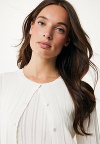 Biała sweterkowa kardigan z pionowymi paskami, z falbaniastym dekoltem i okrągłymi guzikami. Gładka textura z otwartymi detalami dzianiny.