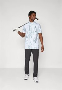 Homem em pé segurando um taco de golfe sobre o ombro, vestindo uma camisa polo estampada, calças pretas, luva de golfe e ténis brancos contra um fundo simples.