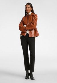 Giacca in ecopelle marrone con bottoni frontali, grande colletto e vestibilità ampia, abbinata a pantaloni neri e stivali alla caviglia. Design pulito e minimalista.