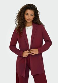 Blazer long bordeaux en tissu doux, avec un col classique, une ouverture devant et des manches longues, associé à un haut blanc en dessous.