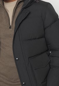 Zbliżenie osoby noszącej brązowy sweter zapinany na zamek pod czarną kurtką puchową z kieszenią na patkę zapinaną na guziki oraz widocznym zamkiem błyskawicznym.