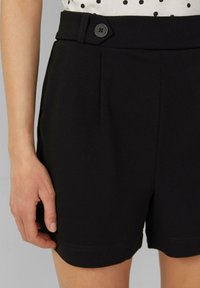 Bermudas negras ajustadas que presentan bolsillos laterales, un cierre de botón visible y pliegues en la parte delantera. Confeccionadas en una tela suave con un acabado limpio.