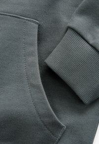 Felpa grigia con tasca frontale e polsini a costine. La texture è liscia, con dettagli di cucitura ben visibili.