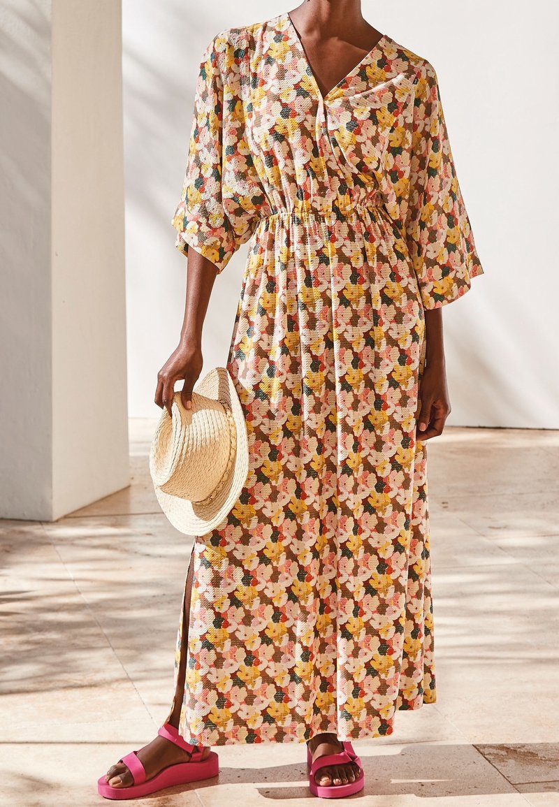 Personne portant une longue robe à motifs floraux avec des manches 3/4, tenant un chapeau de paille dans une main et portant des sandales roses.