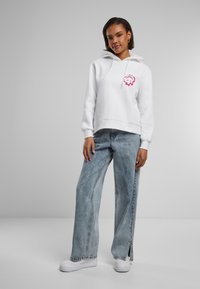 Haut à capuche blanc avec un graphique rose et des cordons de serrage, associé à un jean large bleu clair avec des zips latéraux et des baskets blanches.