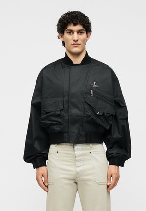 Άνδρας με σγουρά μαλλιά φοράει μαύρο bomber μπουφάν με πολλές τσέπες και μπεζ παντελόνι, στέκεται μπροστά σε απλό λευκό φόντο.