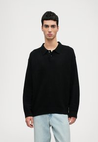Maglione polo nero a maniche lunghe con colletto piatto, realizzato in tessuto morbido. Presenta una vestibilità comoda e tre bottoni sul collo.