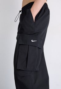 Sorte cargo bukser med snoretræk i taljen, sidelommer og en stor klaftpoche. Har et hvidt Nike-logo på venstre ben.