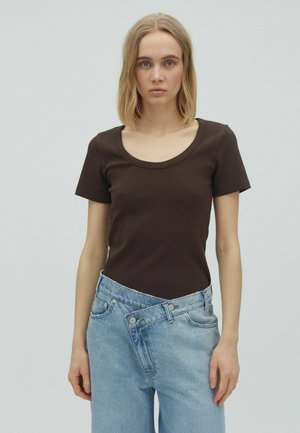 Jeune personne aux cheveux blonds mi-longs portant un haut marron foncé ajusté à manches courtes et un jean bleu clair ample sur un fond uni.