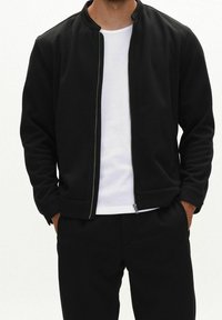 Mann trägt schwarze Jacke mit Reißverschluss über weißem, geripptem T-Shirt, die Hände in den schwarzen Hosentaschen, Bildausschnitt von Hals bis Oberschenkeln.