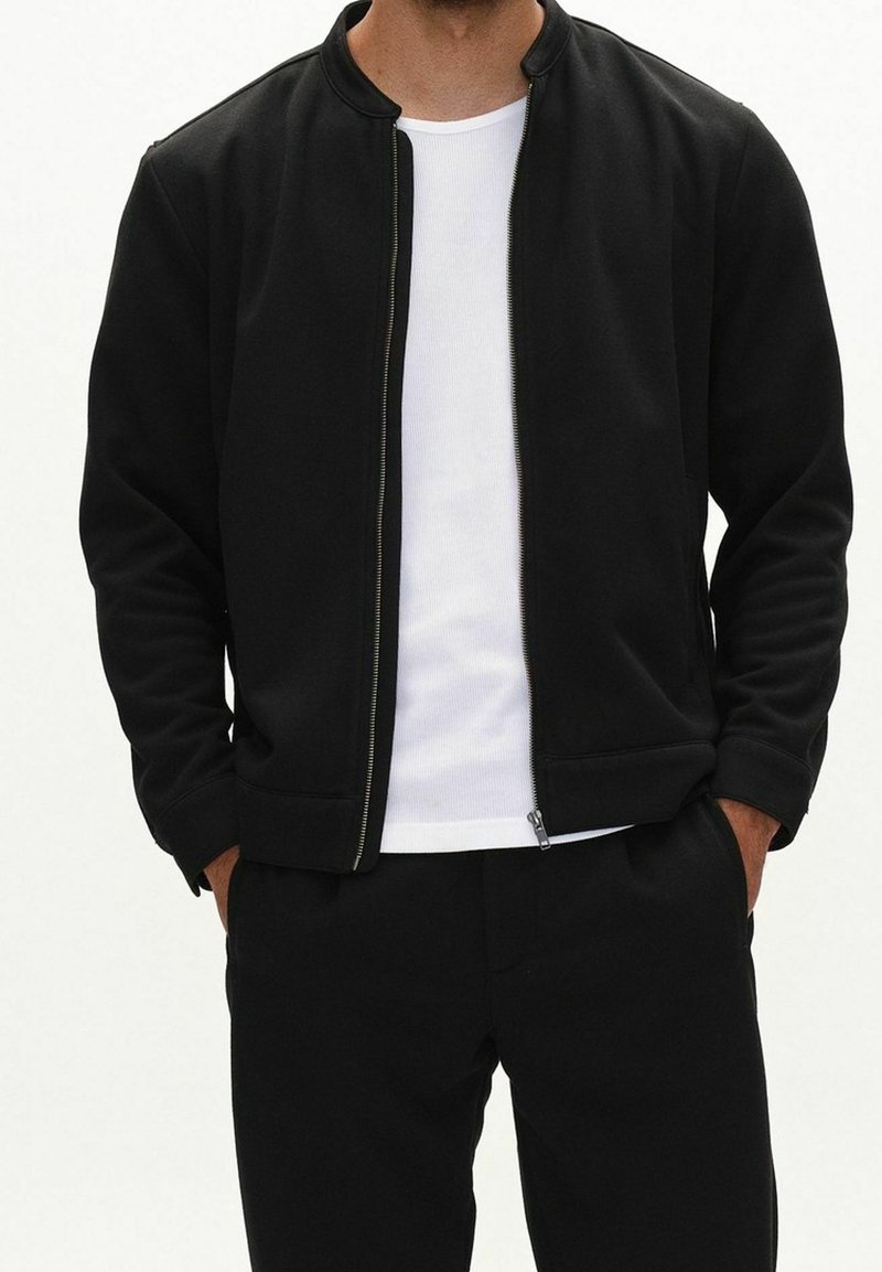 Mann trägt schwarze Jacke mit Reißverschluss über weißem, geripptem T-Shirt, die Hände in den schwarzen Hosentaschen, Bildausschnitt von Hals bis Oberschenkeln.