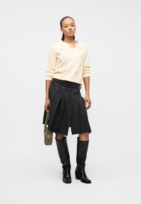 Γυναίκα με πλεξούδες φοράει κρεμ ζακέτα με λουλούδια, μαύρη πλισέ φούστα, μαύρες μπότες μέχρι το γόνατο και κρατά μια τσάντα σε ελιά πράσινο.