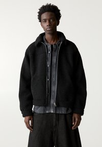 Veste en polaire noire avec fermeture éclair, grand col et surface texturée. Portée par-dessus une chemise boutonnée sombre et un pantalon noir à jambes larges.