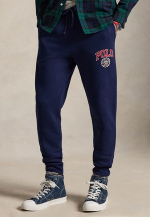Uomo che indossa pantaloni della tuta blu navy con logo rosso "Polo", sneakers alte blu, calzini grigi e camicia verde a quadri.