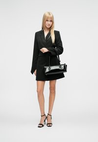 Μαύρο structured blazer φόρεμα με μακριά μανίκια και εφαρμοστή σιλουέτα, συνδυασμένο με γυαλιστερή μαύρη τσάντα χειρός και σανδάλια ψηλοτάκουνα με λουράκια.