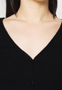 Černý žebrovaný kardigan s výstřihem do V, zdobený řadou černých knoflíků a texturovanou tkaninou. Detailní pohled na oblast krku a ramen.