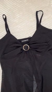 Top preto sem mangas com um detalhe frontal torcido e um acento circular dourado no centro. Feito de um tecido suave e leve com uma textura lisa.
