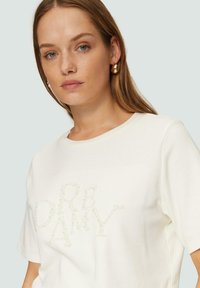 Wit T-shirt met korte mouwen, voorzien van een textuur van kraalversiering die het woord "DREAMY" spelt. Zacht materiaal en een relaxed fit met een ronde halslijn.