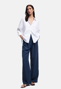 Chemise boutonnée en lin blanc associée à un pantalon large bleu marine, offrant une coupe décontractée et un design simple. Des sandales complètent la tenue.