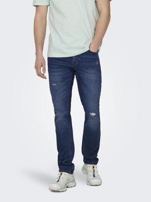 Homme portant un jean déchiré bleu foncé, des baskets blanches et une chemise à manches courtes de couleur claire, une main dans la poche.