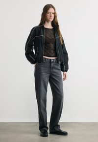 Γυναίκα όρθια που φοράει μαύρο μπουφάν με φερμουάρ, διάφανο μαύρο τοπ με σχέδια, φαρδιά σκούρα γκρι τζιν και μαύρα μοκασίνια μπροστά σε απλό φόντο.