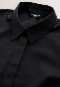 Chemise noire à col pointu avec patte de boutonnage cachée, étiquetée "The Essential Shirt NEXT", posée à plat sur une surface claire.