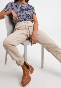 Person in beigen Kordelzug-Jogginghosen, braunen klobigen Stiefeln und einem blauen gemusterten T-Shirt, sitzt auf einem hellgrauen Stuhl mit gespreizten Beinen.