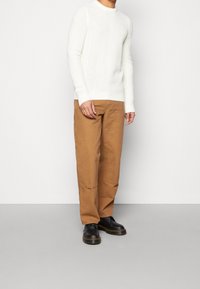 Witte gebreide trui met een gestructureerd patroon, gecombineerd met bruine wijde broek en zwarte veterschoenen. Neutrale achtergrond benadrukt de details van de kleding.